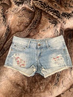 Blå jeansshorts med blommiga detaljer - Säljer ett par ljusblå jeansshorts med råa kanter och slitningar framtill. Under slitningarna syns blommigt tyg i rött, rosa och vitt som ger en unik touch. Klassisk femficksmodell med knapp och dragkedja. Perfekta för sommaren!