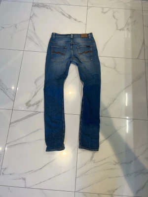 Blå raka Nudie jeans - Snygga blå jeans med raka ben och lätt slitna detaljer. Jeansen har klassisk femficksdesign, kontrastsömmar och läderpatch bak i midjan. Tillverkade i denim med en tvättad look som ger en cool vibe. Osäker på modell