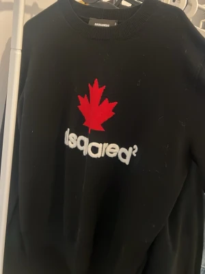 Svart Dsquared2 sweatshirt med lönnlöv - Cool svart sweatshirt från Dsquared2 med ett stort rött lönnlöv och vit broderad logga på bröstet. Tröjan har rund halsringning och är tillverkad i mjukt material, perfekt för en avslappnad streetstyle-look.