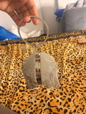 Silver kuvertväska med strass och fransar - Säljer en glittrig silverfärgad kuvertväska täckt av strass och med dekorativa fransar. Väskan har en rund form och ett handtag i metall med strassdetaljer. Perfekt för dig som vill sticka ut och älskar bling!