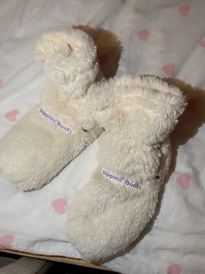 Fluffiga vita tofflor Slippies Boots - Supermysiga vita tofflor från Slippies Boots i fluffigt, mjukt material. De har en hög modell som går upp över ankeln och en brun sula i tyg. Perfekta för att hålla fötterna varma hemma. Broderad logga på sidan. St 38-39 