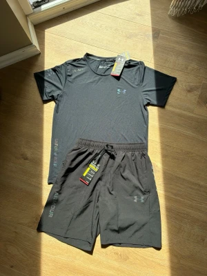 Svart träningsset från Under Armour - Snyggt svart träningsset från Under Armour med t-shirt och shorts. Båda plaggen har diskret logga på bröstet och benet samt tryckt text längs sidan. Materialet är lätt och andas, perfekt för intensiva träningspass. Shortsen har elastisk midja med dragsko.