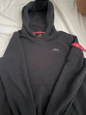 Svart hoodie Alpha Industries - Säljer en svart hoodie från Alpha Industries med stor vit logga på ryggen och mindre logga på bröstet. Hoodien har huva, magficka och en ärmficka med röd tag. Perfekt för dig som gillar streetwear och snygga detaljer.