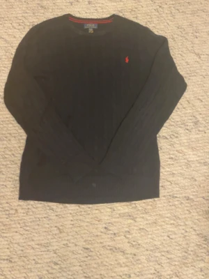 Kabelstickad tröja Polo Ralph Lauren - Klassisk svart stickad tröja från Polo Ralph Lauren med röd broderad logga på bröstet. Storleken är 18-20 år vilket motsvarar S-M. Nypris för denna tröjan är 1800 kr. Tröjan har rund halsringning, ribbade muddar och lång ärm. Perfekt för dig som gillar stilrena och tidlösa plagg. Hör av dig med funderingar eller prisförslag 🤩