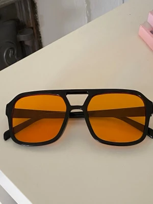 Svarta solglasögon med orange glas - Coola solglasögon med svart, kantig båge och stora orangefärgade glas. De har en modern, sportig design med markerad näsbrygga och breda skalmar.