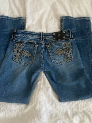 Bootcut Miss Me jeans med broderad ficka - Snygga blå jeans från Miss Me i bootcut storlek 27, midjemått 40cm innerbensläng 78cm. Notera 4de bilden då jeansen har en fläck.