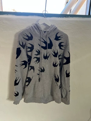 Grå hoodie med svalor från Alexander McQueen  - Snygg grå hoodie från McQ med svarta svaltryck över hela plagget. Klassisk huva med dragsko och känguruficka framtill. Mjuk bomullsblandning som är skön att ha på sig. Perfekt för dig som gillar streetwear och unika prints.