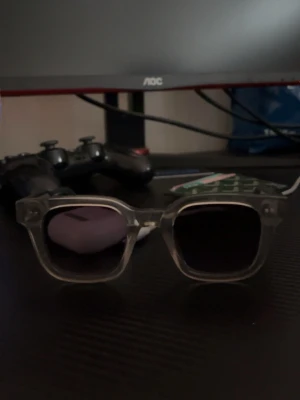 Transparenta solglasögon från Chimi - Snygga solglasögon från Chimi med transparent, fyrkantig båge och mörka glas. Perfekt accessoar för att lyfta din stil med en modern och trendig look. Klassisk design som passar till många outfits.