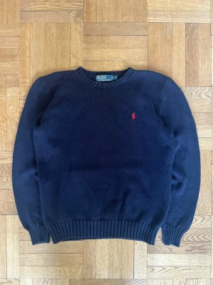Mörkblå stickad tröja Polo Ralph Lauren - Klassisk mörkblå stickad tröja från Polo Ralph Lauren med rund halsringning och ribbade muddar. Tröjan har den ikoniska röda logotypen broderad på bröstet. Perfekt för lager på lager och enkel att matcha med jeans eller chinos.