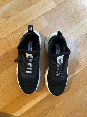 Svarta Steve Madden sneakers med vit sula - Snygga svarta sneakers från Steve Madden med vit chunky sula och svart ovandel i mesh säljes i fint skick . Skorna har elastiska snören med snabblås och tydliga logotyper på plösen och hälen. Perfekta för en trendig och sportig look. Storlek 37, innermåttet är 23 cm.