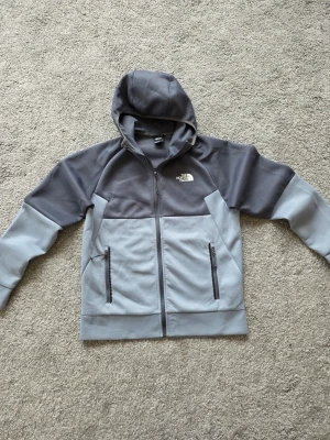 Grå/ Blå hoodie från The North Face - Snygg tvåtonad grå hoodie från The North Face med dragkedja framtill och två praktiska fickor med dragkedja. Hoodien har en klassisk huva och är tillverkad i mjukt fleecematerial, perfekt för chill eller utomhusaktiviteter. Logga på bröstet.