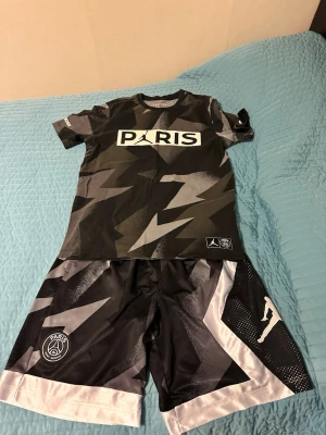 PSG x Jordan svart sportset - Snyggt sportset med t-shirt och shorts från Jordan x PSG. Svart och grått blixtmönster, vita detaljer och loggor på både tröja och shorts. T-shirten har PARIS-tryck framtill och shorts med breda vita kanter. Perfekt för träning eller streetwear. Shortsen är L