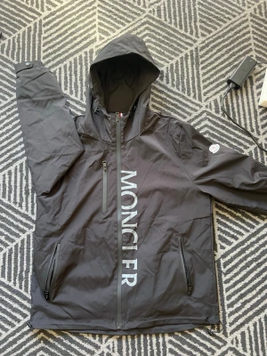 Svart Moncler vindjacka med huva - Svart vindjacka från Moncler med stor logotyp framtill, huva och dragkedja. Jackan har tre fickor med dragkedja och Moncler-märke på ärmen. Materialet är lätt och vattenavvisande, perfekt för blåsiga dagar.