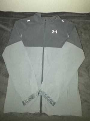Grå vindjacka från Under Armour - Snygg tvåfärgad grå vindjacka från Under Armour med dragkedja framtill och logga på bröstet. Jackan har hög krage, långa ärmar och elastiska muddar med textdetaljer. Perfekt för blåsiga dagar och sportiga outfits.