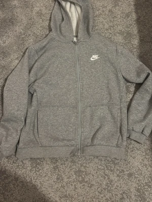 Grå zip-hoodie från Nike - Grå hoodie med dragkedja från Nike. Klassisk modell med huva, två stora fickor framtill och Nike-logga i vitt på bröstet. Mjuk och bekväm, perfekt för en avslappnad stil.