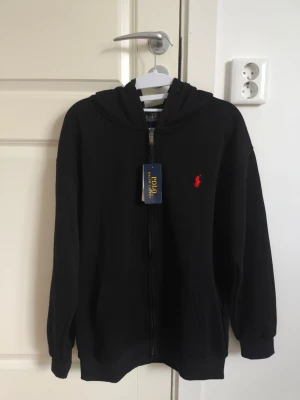 Polo Ralph Lauren Zip Hoodie Svart - Helt ny med tags - Säljer en stilren och klassisk tröja med dragkedja från Polo Ralph Lauren. Tröjan är i nyskick, aldrig använd — säljer eftersom storleken inte passar. Den har en bekväm huva med två dragsnören samt fickor framtill i mitten, vilket gör den både praktisk och skön att använda.      Mått (ca): Axelbredd: 53 cm Längd: 65 cm.           Logotyp: Klassisk broderad logotyp i rött på bröstet. Material: Mjuk och högkvalitativ bomullsblandning med fleece inuti.