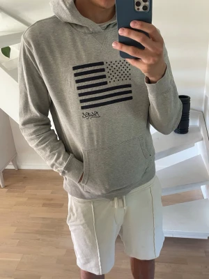 Grå hoodie med USA-flagga från Ralph Lauren - Snygg grå hoodie från Ralph Lauren med mörkblå USA-flagga tryckt på bröstet och signatur under. Klassisk känguruficka framtill och huva. Sitter som Medium.