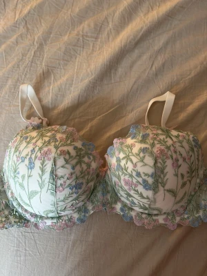 Blommig broderad bh med pastellfärger - Romantisk bh med broderade blommor i pastellfärger på vit botten. Kuporna är vadderade och har spetskant samt en liten rosett mellan kuporna. Axelbanden är vita och justerbara. Perfekt för dig som gillar en feminin och söt stil.