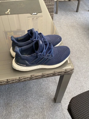 Adidas Ultra Boost marinblå sneakers - Snygga Adidas Ultra Boost sneakers i marinblått med stickad ovandel och klassiska tre ränder på sidan. Skorna har en vit Boost-sula för extra komfort och energiåtergivning. Perfekta för dig som gillar sportig stil och vill ha riktigt sköna sneakers.