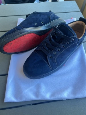 Christian Louboutin - Snygga mörkblå sneakers i mocka med mörkblåa snören och detaljer. Skorna har en unik röd sula och diskret design som passar till många outfits. Insidan är ljusbeige och skon har en klassisk låg profil. Perfekta för dig som gillar stilrena och trendiga sneakers.