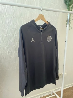 Svart träningsjacka PSG x Jordan - Svart långärmad träningsjacka med halv dragkedja framtill, PSG-logga och Jordan-logga på bröstet. Materialet är syntetiskt och har en sportig känsla, perfekt för träning eller fotboll. Jackan har en modern passform och diskreta detaljer på axlarna.