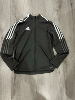 Svart Adidas träningsjacka 164 - Snygg svart träningsjacka från Adidas med vita ränder längs ärmarna och dragkedja framtill. Tillverkad i Primegreen-material som är återvunnet. Jackan har hög krage och Adidas-logga på bröstet. Perfekt för träning eller sportiga dagar.