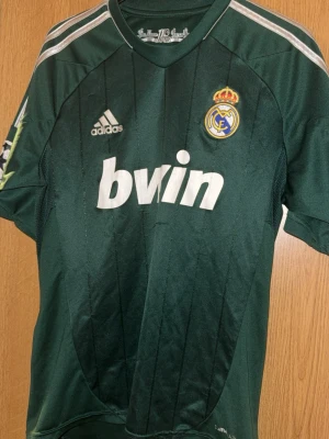 Real Madrid bortatröja Adidas grön - Grön Real Madrid bortatröja från Adidas med klubbmärke och Bwin-logga på bröstet. Tröjan har vita detaljer, korta ärmar och är tillverkad i ClimaCool-material som andas. Perfekt för fotbollsträning eller att bära som supporter.