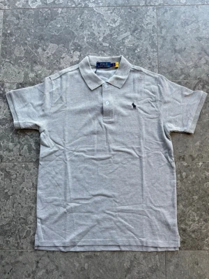 Grå pikétröja från Polo Ralph Lauren - Klassisk grå pikétröja från Polo Ralph Lauren med korta ärmar och krage. Tröjan har två knappar framtill och den ikoniska broderade loggan på bröstet. Tillverkad i mjuk bomull för en bekväm känsla. Viktigt att veta är att den är liten i storleken, passar även dom som vanligtvis har M!