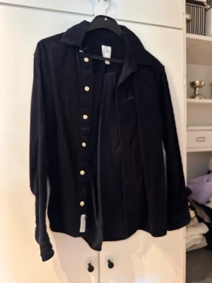 Svart manchester-skjorta med knappar - Snygg svart skjorta i manchester med klassisk krage och vita knappar framtill. Skjortan har lång ärm och en bröstficka. Perfekt för dig som gillar en stilren och trendig look.
