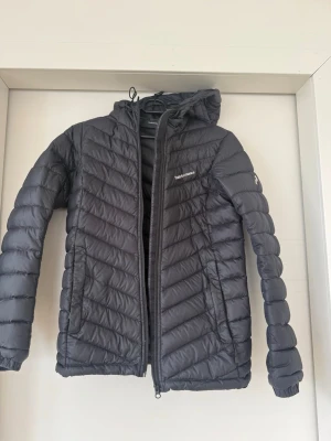 Svart dunjacka Peak Performance - Snygg svart dunjacka från Peak Performance med huva och dragkedja framtill. Jackan har quiltad design och är lätt vadderad för extra värme. Perfekt för kalla vinterdagar och har en stilren look med diskret logga på bröstet.