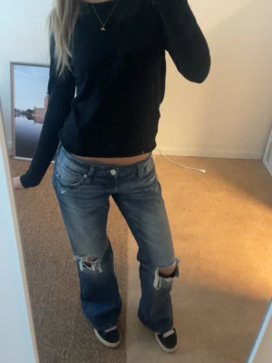 Blå slitna bootcut jeans - Säljer ett par blå bootcut jeans med slitningar och hål på båda benen. Jeansen har låg midja, fem fickor och en avslappnad passform. Perfekta för en avslappnad och trendig look.