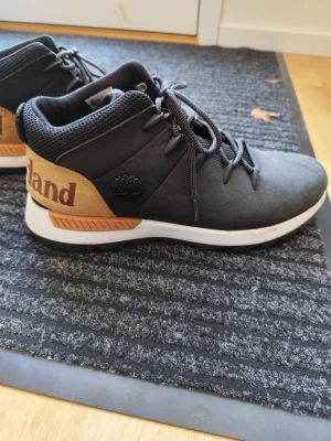Timberland svarta och bruna sneakers - Snygga svarta sneakers från Timberland med bruna detaljer och stor logga på hälen. Skorna har vita sulor, svarta snören och är tillverkade i en mix av läder och textil. Perfekta för dig som gillar streetstyle och vill sticka ut med en klassisk look.