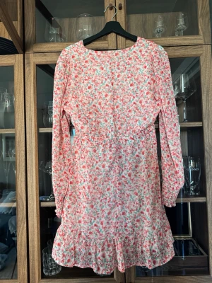 Blommig kortklänning med volang - Fin kortklänning med rosa och röda blommor på vit botten. Klänningen har v-ringning, långa ärmar med resår vid ärmslut och volangkant nertill. Perfekt för vår och sommaren. Klänningen är ifrån Amisu!🤍