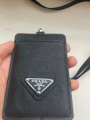 Svart korthållare  - Snygg svart korthållare i tåligt läder med klassisk trekantig logotyp i metall framtill. Kommer med en matchande rem att hänga runt halsen. Perfekt för kort och ID när du vill ha det enkelt och stilrent.