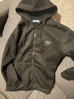 Svart hoodie med dragkedja Prada - Svart hoodie från Prada med dragkedja framtill, huva med snören och två stora fickor. Materialet är mjukt och bekvämt, perfekt för chill dagar. Diskret logotyp på bröstet. Klassisk och stilren design som funkar till det mesta.