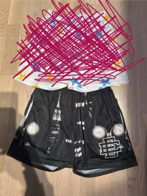 EE Shorts - Svarta shorts i meshmaterial med tryckt stadsmotiv och vit logga på ena benet. Elastisk midja för skön passform. Perfekta för en avslappnad streetwear-look. BARA DEM SVARTA EE SHORTSEN ÄR KVAR 