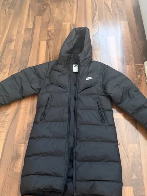 Svart lång dunjacka från Nike - Snygg svart lång dunjacka från Nike med huva och dragkedja hela vägen. Jackan har två snedställda fickor med dragkedja och diskret Nike-logga på bröstet. Perfekt för kalla vinterdagar och ger en sportig look. Skicket är ungefär 7/10, jackan sitter perfekt för alla mellan 170-175 den kan sitta lite olika på alla men sitter väldigt bra för smala personer. Rivning i inre jackan kolla sista bilden