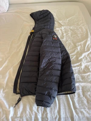 Svart dunjacka med huva från Parajumpers - Säljer en svart dunjacka från Parajumpers med huva och gul detalj vid halsen. Jackan har quiltad design, dragkedja och patch på ärmen. Insidan är ljusgrå och jackan är tillverkad i ett lätt och varmt material som passar perfekt för kalla vinterdagar.