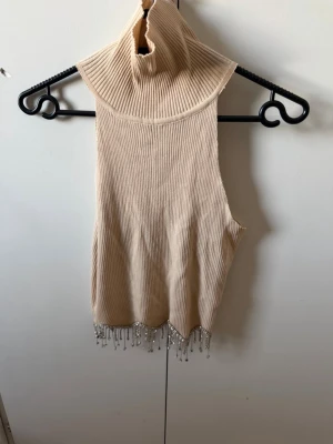 Beige ribbad polotopp med pärlor - Trendig beige ribbad polotopp från Giorgia med ärmlös design och snygga pärldetaljer längs nederkanten. Perfekt för dig som vill sticka ut med en unik topp som har hög krage och coolt fall.