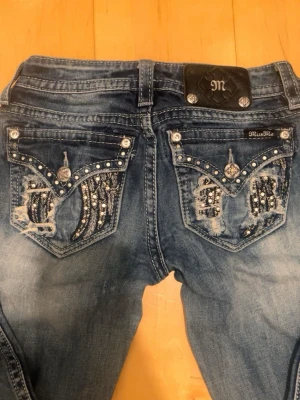 Miss Me Jeans - Snygga bootcut jeans från Miss Me med dekorativa bakfickor med paljetter, nitar och broderier. Jeansen har en mycket fin färg! Jeansen har låg midja, klassisk femficksmodell och silverfärgade knappar. Har en fläck som inte går bort från förra ägaren! Motsvarar ungefär en storlek M/S