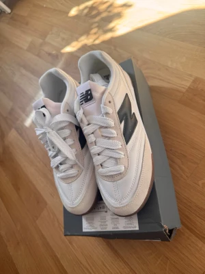 Vita New Balance sneakers med svart logo ,helt nya - Snygga vita New Balance sneakers med svart N-logga på sidan och klassisk retrostil. Ovandelen är i läder och mocka, med vita skosnören och brun sula. Perfekta för dig som gillar stilrena och sportiga skor med en tidlös vibe.