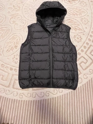 Svart dunväst med huva - Snygg svart dunväst med huva och dragkedja framtill. Västen har quiltad design och är perfekt för lager-på-lager-stil. Passar dig som gillar en clean och sportig look. Materialet är glansigt och lätt, vilket gör den smidig att bära.