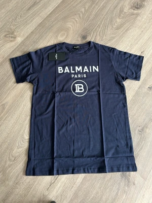 Mörkblå Balmain Paris t-shirt - Snygg mörkblå t-shirt från Balmain Paris med vit logga och tryck framtill. Klassisk passform med rund hals och korta ärmar. Perfekt för dig som gillar exklusiva streetwear-plagg.