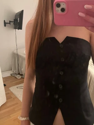 Svart axelbandslös korsettopp - Snygg svart axelbandslös korsettopp med knappar framtill och en liten slits nedtill. Toppen har en stilren och elegant design som passar perfekt till många olika outfits. Materialet känns mjukt och följsamt mot kroppen.