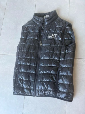 Svart dunväst från Emporio Armani EA7 - Snygg svart dunväst från Emporio Armani EA7 med quiltad design och hög krage. Västen har dragkedja framtill och logotyp på bröstet. Perfekt för lager-på-lager och ger en cool look. Materialet är glansigt och lätt, vilket gör den enkel att bära.