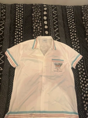 Casablanca Tennis Club skjorta - Vit kortärmad skjorta från Casablanca med pastellblå och rosa ränder på krage, ärmslut och nederkant. Bröstficka med broderad tennisklubbslogga. Perfekt för en sportig och trendig look.