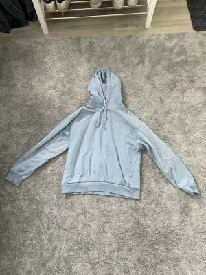 Ljusblå hoodie med huva - En stilren ljusblå hoodie med huva och dragsnören. Hoodien har ribbade muddar vid ärmslut och nederkant samt en klassisk passform. Perfekt för en avslappnad look. Materialet känns mjukt och bekvämt mot huden. Snörerna är slute upp snurrade.