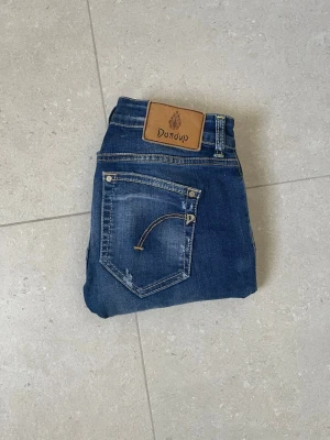 Blå jeans från Dondup med slitningar - Säljer ett par blå jeans från Dondup med snygga slitningar och kontrastsömmar. Jeansen har klassisk femficksmodell, läderpatch med logga bak och raka ben. Materialet är denim i bomull med en cool tvättad look. Passar folk runt 160cm+. Säljer dem på grund av att jag tycker att de är lite tajta runt låren. Men är annars mycket bra skick.