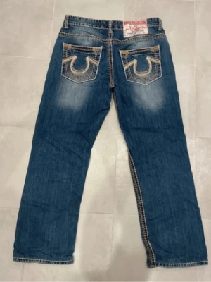True Religion blå jeans med kontrastsöm - Säljer ett par jättefina  blå jeans från True Religion. De är köpta av mig innan här på Plick och är knappt använda av mig. Det finns ett lagat hål precis över vänstra bakfickan (bild 4) men annars är de i fint skick! Det står strl 33 och passar mig med lite marginal som brukar ha strl 40. Jag är 170 och de nuddar inte marken.