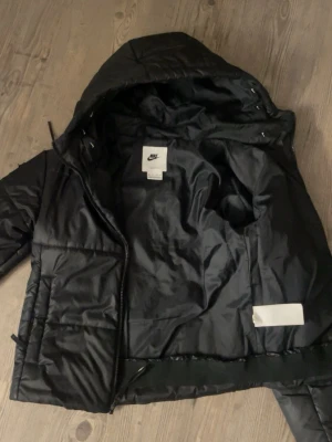Svart pufferjacka från Nike - Säljer en svart pufferjacka från Nike med huva och dragkedja framtill. Jackan har två fickor på sidorna och en vit Nike-logga på bröstet. Tillverkad i ett glansigt material som håller värmen under kalla dagar. Perfekt för dig som vill ha en stilren och sportig look. 🔥